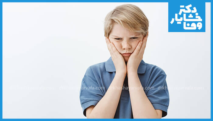 پسر بچه‌ای با موهای روشن و پیراهن آبی که هر دو دستش را روی گونه‌ها گذاشته و حالت چهره‌اش نشان‌دهنده درد است؛دندان‌درد دارد.