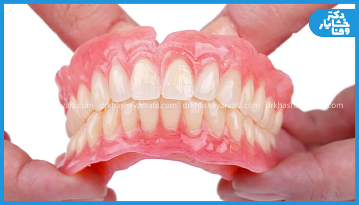 یک عکس کلوزآپ و باکیفیت از یک دست دندان مصنوعی که توسط دو دست نگه داشته شده است، و هر دو فک بالا و پایین را نشان می‌دهد. دندان‌های مصنوعی برای شبیه‌سازی لثه صورتی و برای دندان‌ها سفید هستند.