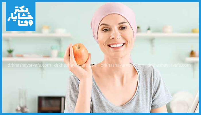 زن مبتلا به سرطان با کلاه سر صورتی، در حال لبخند زدن و نگه‌ داشتن یک سیب، تأکید بر سلامت دهان در طول شیمی درمانی.